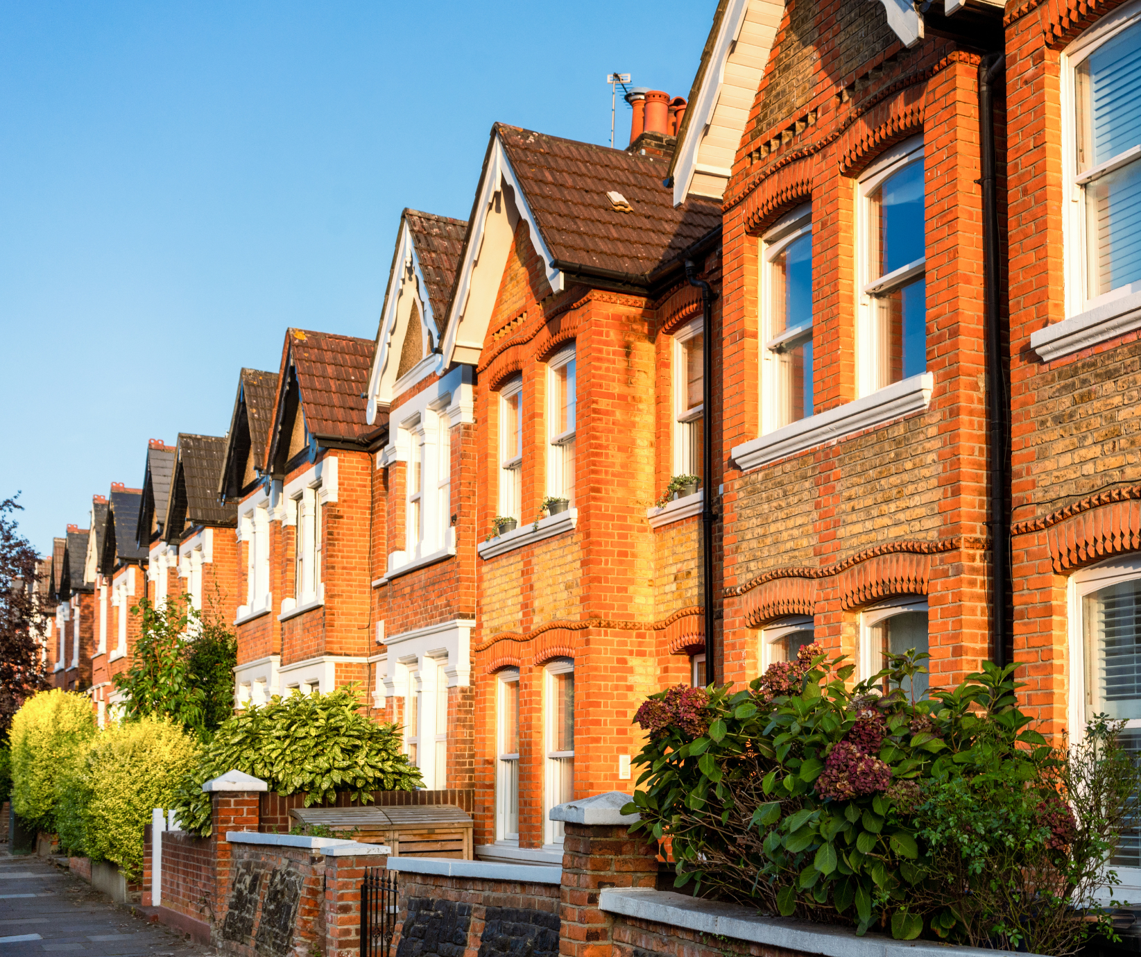 A row of attached red-brick terraced houses with bay windows and pitched roofs, lined along a residential street with front gardens and shrubs, illuminated by warm sunlight under a clear blue sky.