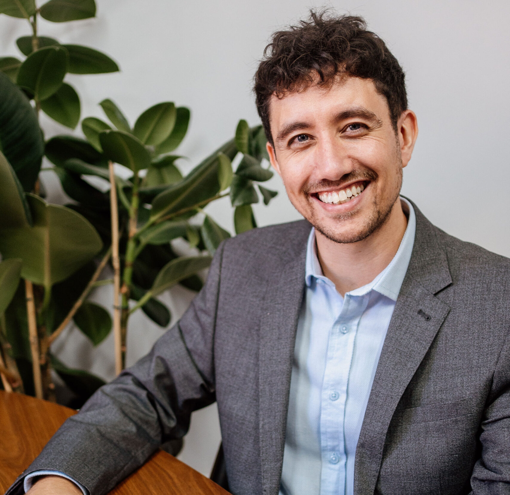 Martyn King, a smiling man in a grey blazer and light blue shirt, sitting at a wooden table with a document and pen, with green houseplants in the background.